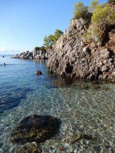 Clear water with some large rocks, a little drop off, someone swimming in the distance a bit and a rock land formation on the right side.